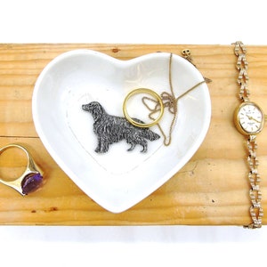 May include: A white heart-shaped ceramic dish with a silver dog figurine, a gold necklace, and a gold ring with a purple stone. The dish is on a wooden surface with a gold watch with a white face.