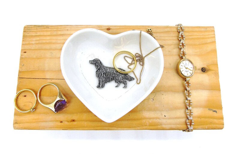 May include: A white heart-shaped ceramic dish with a silver dog figurine, a gold necklace, and a gold ring with a purple stone. The dish is on a wooden surface with a gold watch with a white face.