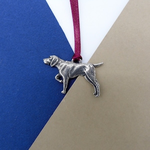 Może przedstawiać: Srebrny, cynowy ornament w kształcie psa wiszący na burgundowym sznurku. Pies stoi z głową zwróconą w lewo.