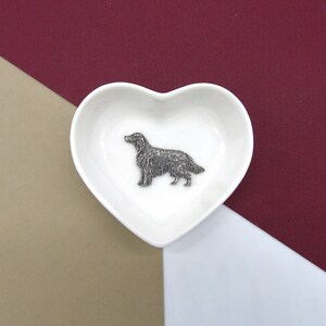 May include: A white heart-shaped ceramic dish with a silver metal figurine of a dog inside. The dish is on a brown and burgundy background.