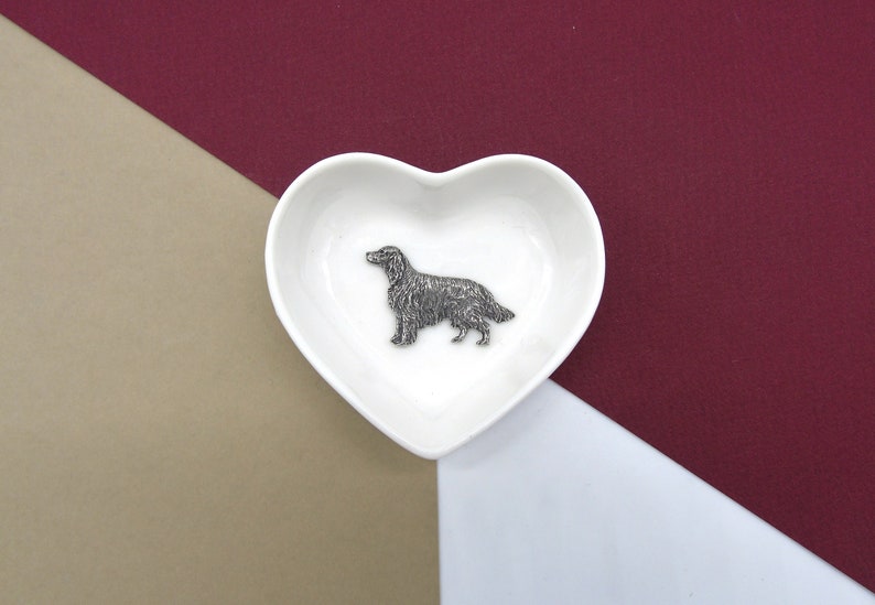 May include: A white heart-shaped ceramic dish with a silver metal figurine of a dog inside. The dish is on a brown and burgundy background.