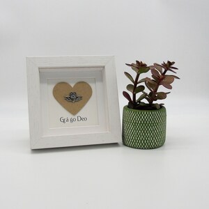 May include: A white frame with a brown heart and a silver Claddagh charm inside. The frame is sitting on a white surface next to a small green potted plant. The text "Gr&aacute; go Deo" is written below the heart.