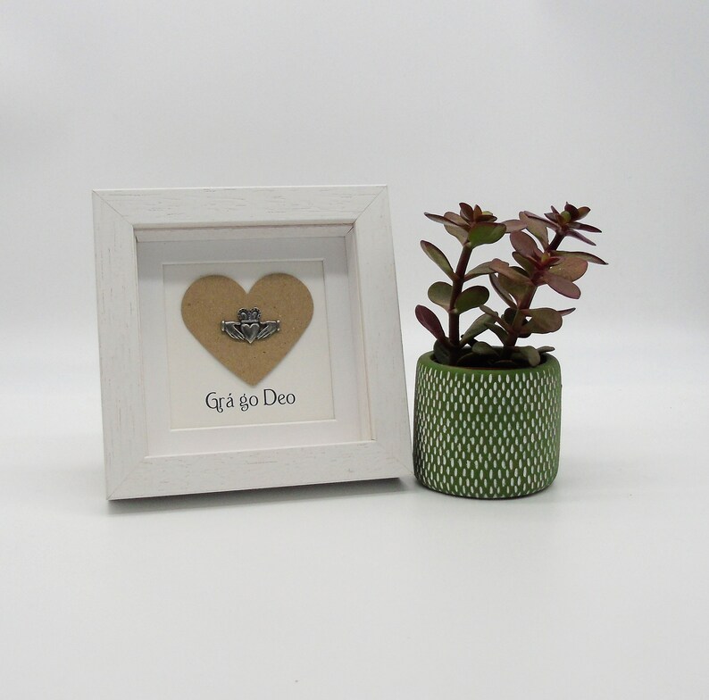 May include: A white frame with a brown heart and a silver Claddagh charm inside. The frame is sitting on a white surface next to a small green potted plant. The text "Gr&aacute; go Deo" is written below the heart.