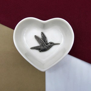 May include: A white heart-shaped dish with a silver hummingbird figurine inside.