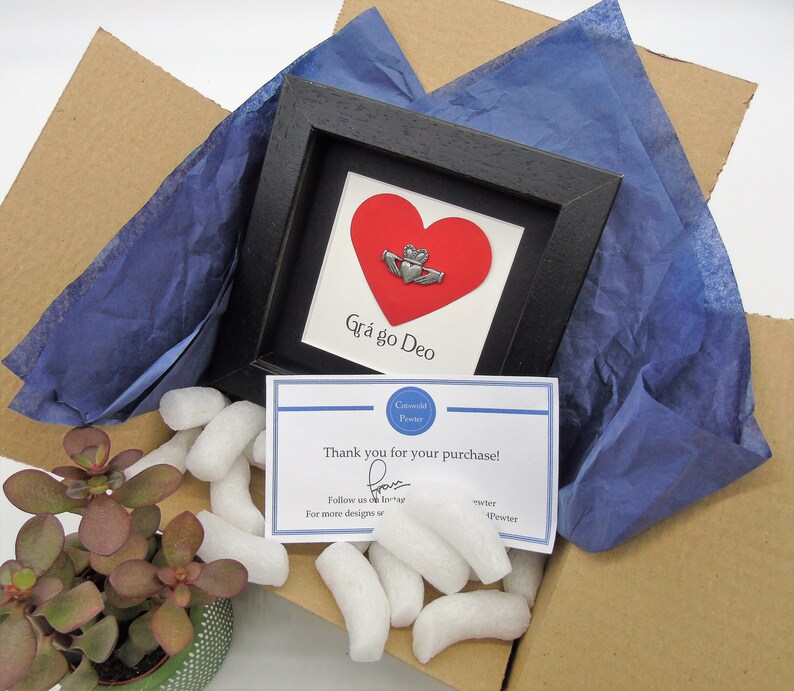 May include: A small framed print with a red heart and a silver Celtic knot. The text "Gr&aacute; go Deo" is written below the heart. The print is packaged in a cardboard box with blue tissue paper and white packing peanuts.