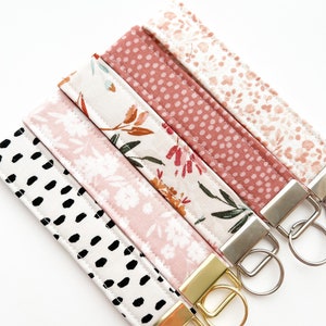 May include: Set of six keychains with pink and white fabric straps and gold and silver key rings. The straps feature different patterns including floral, polka dots, and abstract designs.