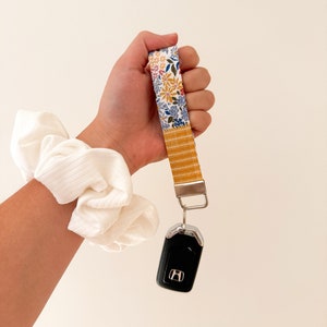 May include: A floral patterned key fob wristlet with a gold-colored stripe and a silver-colored key ring holding a black car key. A white scrunchie is on the wrist. The wristlet features a colorful floral design with blue, yellow, and red flowers.