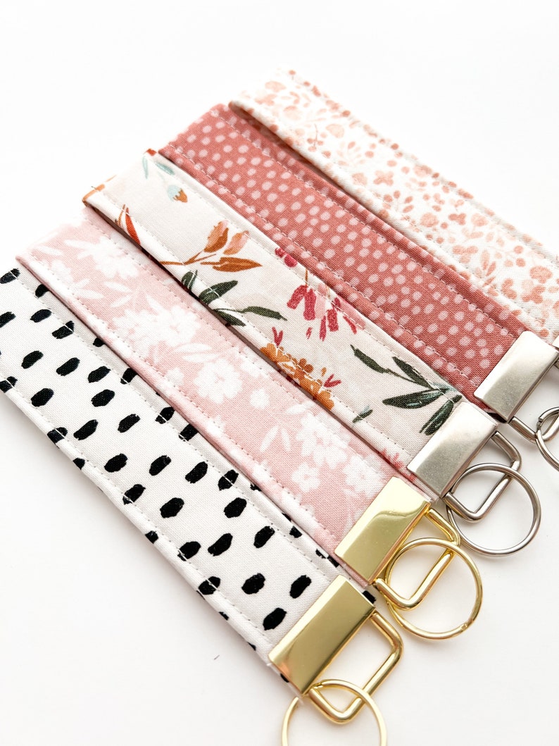 May include: Five keychains with different fabric patterns. The keychains are pink, white, and black with floral and polka dot designs. Each keychain has a gold or silver key ring.