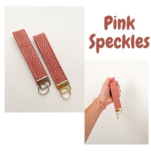 May include: Two pink keychains with white polka dots and gold key rings. The keychains are made of fabric and have a simple design. The text "Pink Speckles" is visible in the image.