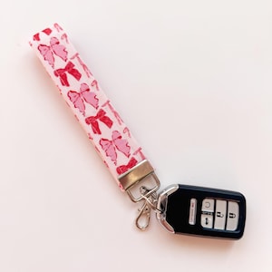 May include: A pink and red key chain with a silver clasp and a black car key fob. The key chain has a pattern of red bows on a pink background.