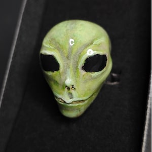 Könnte beinhalten: Eine grüne Alien-Kopfskulptur mit schwarzen Augen und einem kleinen Mund. Die Skulptur hat eine glatte, glänzende Oberfläche und befindet sich vor einem schwarzen Hintergrund.