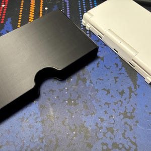 May include: A matte black rectangular plastic case with a curved cutout on one side, positioned next to a closed white handheld gaming console. The console features a hinge mechanism and visible ports along the side edge of its outer shell.