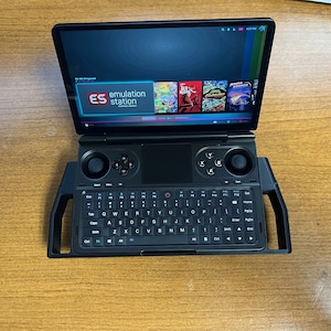 Op de afbeelding: Een zwart draagbaar gameapparaat met een ingebouwd toetsenbord en twee analoge joysticks. Het scherm toont het logo "ES emulation station" en verschillende game-pictogrammen.