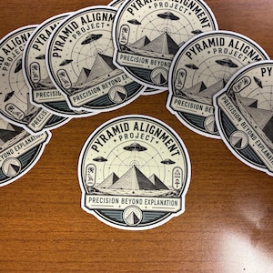May include: A collection of circular stickers featuring a graphic illustration of pyramids under flying saucers. The text reads "PYRAMID ALIGNMENT PROJECT" and "PRECISION BEYOND EXPLANATION," accompanied by Egyptian hieroglyphics and a grid pattern.