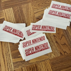 May include: Several white stickers featuring the text "SUPER NINTENDO ENTERTAINMENT SYSTEM" in red. The stickers are stacked on a wooden surface with a parquet design. The text is in a bold, sans-serif font.