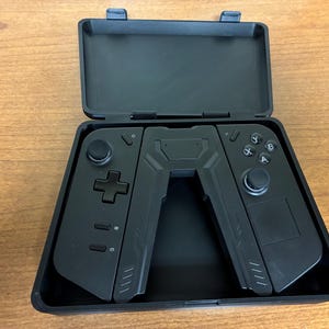 May include: Black gaming controller in an open black case. The controller is split into two halves, each with buttons, joysticks, and a directional pad. The case is rectangular with a hinged lid.