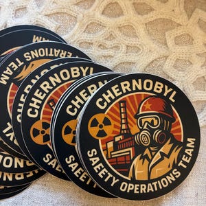 Op de afbeelding: Een stapel ronde zwarte stickers met een retro-ontwerp. De stickers tonen het woord "Chernobyl" in een cirkelvormig patroon, samen met een afbeelding van een persoon met een gasmasker en de tekst "Safety Operations Team."