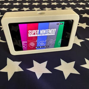 May include: A white rectangular smartphone stand holding a phone displaying a Super Nintendo Entertainment System interface with colourful vertical panels and controller icons. The stand features a smooth finish and a circular cutout on the top edge.