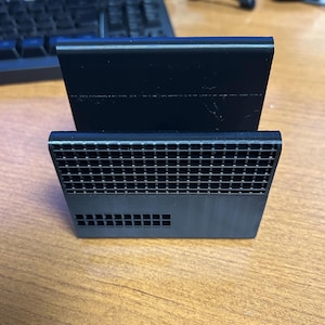 Puede incluir: Un soporte de plástico negro con un patrón de rejilla en la parte delantera y una superficie lisa en la parte superior. El soporte está diseñado para sostener un dispositivo en posición vertical.