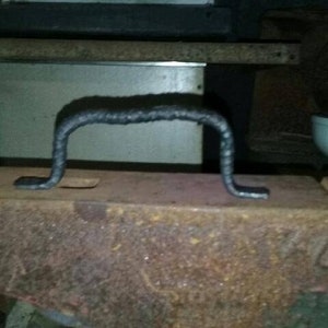 May include: A black metal handle with a rounded top and a flat base. The handle is sitting on a rusty metal surface.