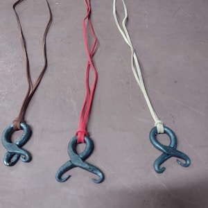May include: Three handmade metal pendant necklaces. Each necklace features a dark gray, abstract metal pendant on a leather or suede cord. The cords are brown, red, and cream-colored, and are attached to the pendant with a silver clasp.