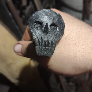 May include: A black metal skull head with a detailed design. The skull has two eye sockets, a nose, and a mouth with teeth.