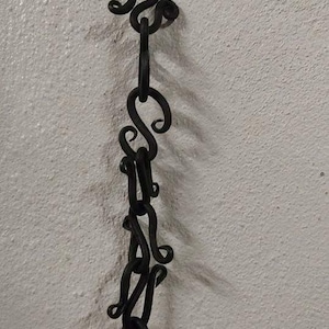 Może przedstawiać: Czarny metalowy łańcuch z wieloma ozdobnymi pętlami. Łańcuch wisi na białej ścianie.