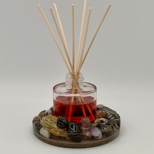 May include: A glass diffuser bottle with red liquid and wooden sticks, sitting on a round base decorated with various stones and shells.