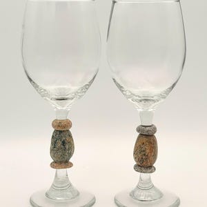 May include: Two clear wine glasses with unique stems. The stems feature natural stone accents in shades of brown, gray, and beige. The glasses are designed for serving wine and add a rustic touch to any table setting.