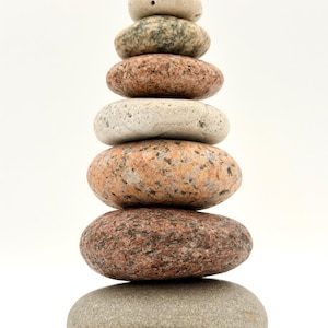 May include: A stack of seven smooth, rounded stones of varying sizes and colors, balanced on top of each other. The stones range in color from light gray to reddish-brown, creating a natural, minimalist sculpture against a white background.