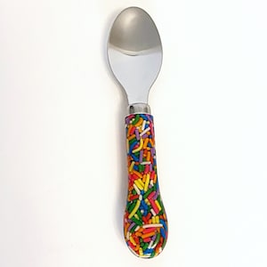 May include: A silver spoon with a colorful sprinkle-patterned handle. The handle is made of a clear resin with embedded sprinkles in various colors, including red, orange, yellow, green, blue, and purple.