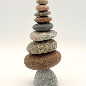 May include: A vertical stack of nine smooth, rounded river stones balanced on top of each other. The stones vary in size and color, ranging from light gray and speckled granite to dark charcoal and reddish-brown hues, creating a tapered tower shape.