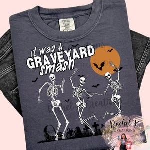 May include: Dark blue t-shirt with the text "it was a GRAVEYARD smash" in white. The shirt features a Halloween design with dancing skeletons, bats, and an orange moon. A tombstone and pumpkins are also visible.