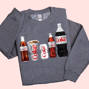 May include: A gray sweatshirt featuring a graphic of Diet Coke bottles, cans, and a cup. The Diet Coke logo is prominently displayed on each item. The sweatshirt has a crew neck and long sleeves. The brand name "Gildan" is visible on the tag.