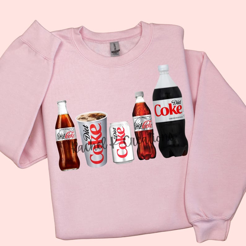 May include: A light pink sweatshirt featuring a Diet Coke design. The design includes images of Diet Coke bottles, a can, and a cup. The text "Diet Coke" is visible on the items. The sweatshirt has long sleeves and a crew neck.