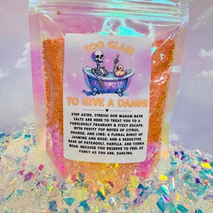 Puede incluir: Una bolsa de plástico transparente llena de sales de baño de color naranja. La bolsa tiene una etiqueta que dice "Too Glam To Give A Damn!" con un esqueleto de dibujos animados en una bañera. La etiqueta también describe las sales de baño como "fabulosamente fragantes y efervescentes" con notas de cítricos, jazmín, rosa, pachulí, vainilla y haba tonka.