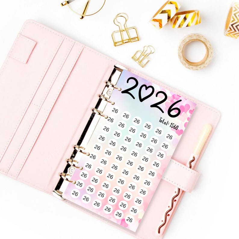 May include: A pink planner open to a page with a design featuring the year "2026" and the number "26" repeated. The text "Total: 1560" is also visible. Gold binder clips, washi tape, and a pen are scattered around the planner.