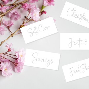May include: Five white cards with handwritten text on a white background. The cards read "Christmas", "Self Care", "Savings", "Food Shop", and "Fant: 3". Pink cherry blossoms are scattered around the cards.