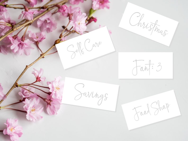 May include: Five white cards with handwritten text on a white background. The cards read "Christmas", "Self Care", "Savings", "Food Shop", and "Fant: 3". Pink cherry blossoms are scattered around the cards.