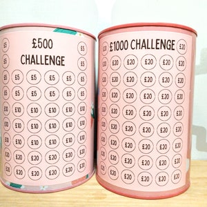 Może przedstawiać: Dwie cylindryczne metalowe puszki na pieniądze z r&oacute;żowymi etykietami. Lewa puszka ma napis &pound;500 CHALLENGE z rzędami k&oacute;łek oznaczonych &pound;5, &pound;10 i &pound;20. Prawa puszka ma napis &pound;1000 CHALLENGE z rzędami k&oacute;łek oznaczonych &pound;20.