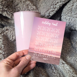 May include: A pink and white card with the text "Holiday Fund" and a grid of 49 white airplane icons on a pink and orange sunset background. The card has a line at the bottom for a total amount.