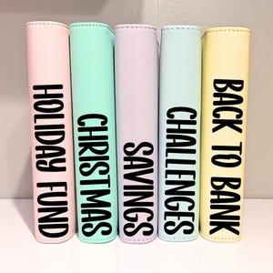 May include: Five vertical binders in pastel colours including pink, mint green, lavender, light blue, and yellow. Each binder features bold black text on the spine reading HOLIDAY FUND, CHRISTMAS, SAVINGS, CHALLENGES, and BACK TO BANK respectively.