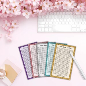May include: Five rectangular Penny Saving Challenge trackers with glittery borders in purple, pink, silver, teal, and gold. Each tracker features a grid of numbers and the text Penny Saving Challenge. A white pen rests on the gold tracker.