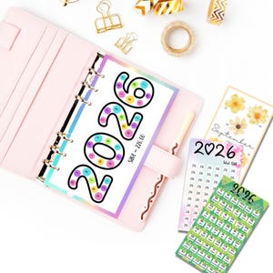 May include: A pink planner with a 2026 savings tracker insert, featuring colorful circles. Additional inserts include a September calendar and a savings tracker. Gold binder clips and decorative tape rolls are also visible.