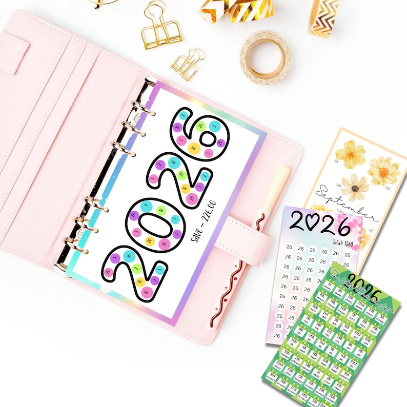 May include: A pink planner with a 2026 savings tracker insert, featuring colorful circles. Additional inserts include a September calendar and a savings tracker. Gold binder clips and decorative tape rolls are also visible.
