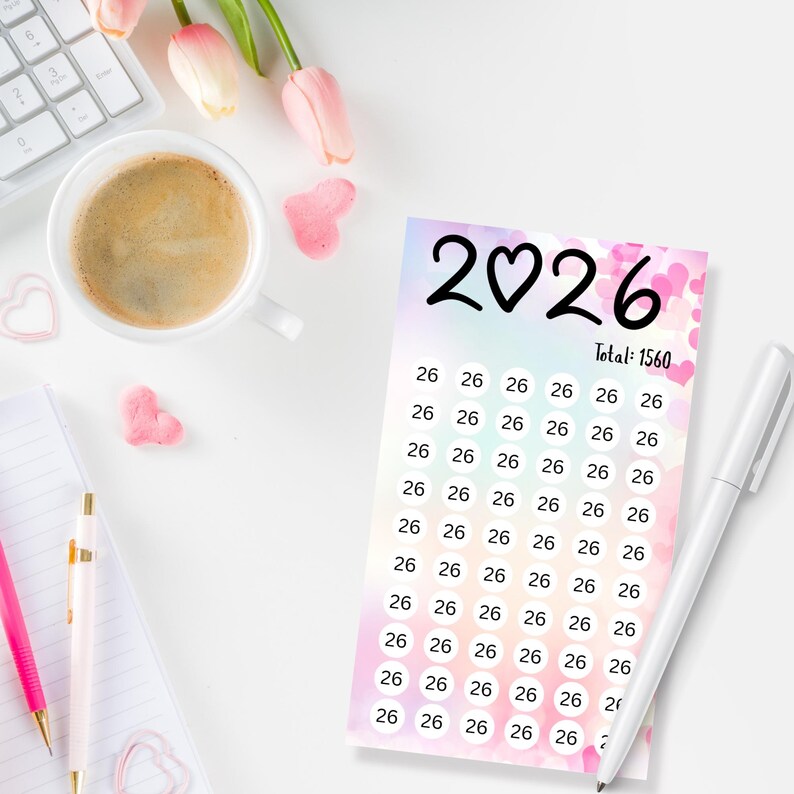 May include: A planner card with the year "2026" and a heart symbol, with the total of "1560". The card has a pastel background with pink hearts and the number "26" repeated. A white pen rests on the card. A cup of coffee, a keyboard, and pink flowers are also visible.