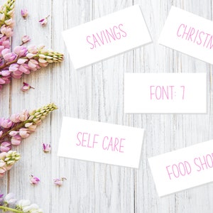 May include: White cards with pink text on a white wooden background. The cards say "Savings", "Christmas", "Food Shop", "Self Care", and "FONT: 7".  Pink flowers are in the left corner of the image.