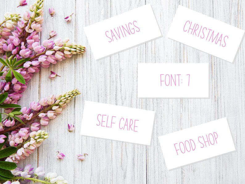 May include: White cards with pink text on a white wooden background. The cards say "Savings", "Christmas", "Food Shop", "Self Care", and "FONT: 7".  Pink flowers are in the left corner of the image.