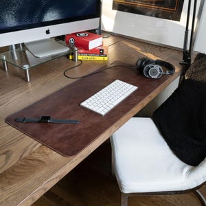 Könnte beinhalten: Eine rechteckige Schreibtischunterlage aus dunkelbraunem Leder mit sichtbaren Nähten an den Rändern, die auf einem Schreibtisch aus Holz liegt. Eine weiße kabellose Tastatur, eine schwarze Smartwatch und ein schwarzer Kopfhörer liegen darauf.