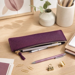 Puede incluir: Un estuche rectangular de color morado con superficie texturizada y cierre de cremallera dorado. El estuche está parcialmente abierto, mostrando un bolígrafo blanco en su interior. Un bolígrafo morado a juego y clips dorados están cerca.